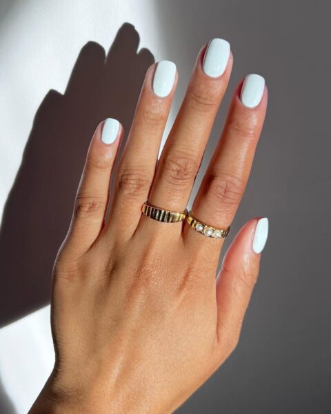 Zdjęcie dłoni ze zbliżeniem na manicure. To modne, białe paznokcie na maj 2025, czyli kwadratowe i w odcieniu kredowej bieli. Na palcach znajdują się złote pierścionki. Dłoń pozuje na tle białej ściany.