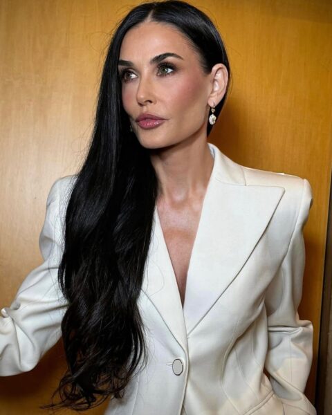 Zdjęcie aktorki Demi Moore w długich, czarnych włosach. Ma mocny makijaż z wyraźnym konturowaniem. Ubrana jest w kremową, taliowaną marynarkę. W uszach wiszące kolczyki z cyrkonią. Pozuje na tle drewnianych drzwi.