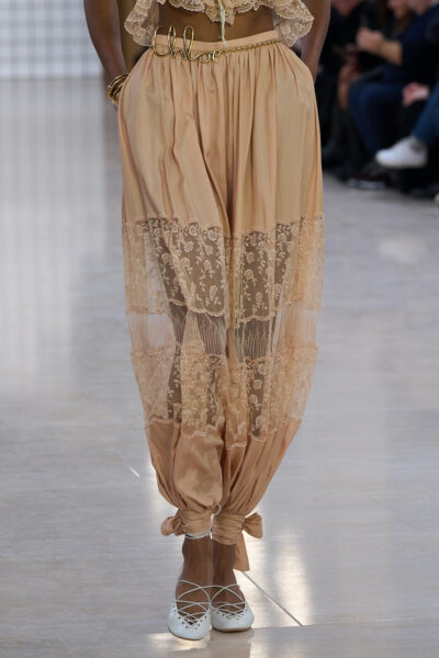 Modelka na wybiegu prezentuje eleganckie spodnie w stylu boho od Chloe. Spodnie mają pastelowy, brzoskwiniowy odcień i są ozdobione koronkowymi, przezroczystymi wstawkami w kwiatowy wzór. W pasie widoczny jest złoty pasek z napisem 'Chloé', a nogawki są zebrane na wysokości kostek i zawiązane w kokardy. Stylizację dopełniają białe baleriny z krzyżującymi się paseczkami. To spodnie na lato, które stylizować można na co dzień, do pracy i na plażę.