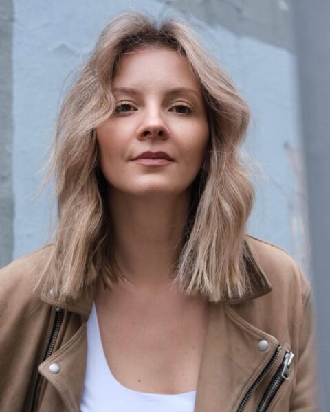 Zdjęcie dojrzałej kobiety w blond włosach do ramion. Są cieniowane, pofalowane. Ma do tego grzywkę na boki. Ubrana jest w biały top oraz beżową kurtkę w skórzaną.