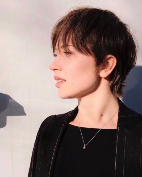 Zdjęcie dojrzałej kobiety w krótkich, ciemnych włosach ściętych w stylu pixie cut. Ubrana jest w czarną bluzkę i czarną marynarkę. Ma na szyi drobny, srebrny łańcuszek z zawieszką. Pozuje półbokiem.
