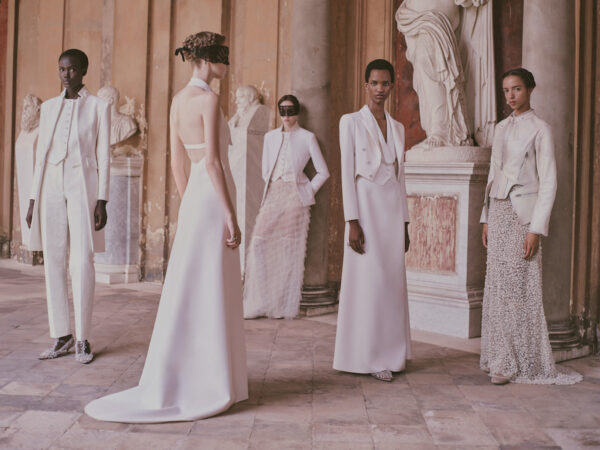 Grupa pięciu modelek pozuje w eleganckim wnętrzu w stylu klasycznym, na tle marmurowych rzeźb i kolumn. Ubrane są w stylizacje haute couture domu mody Dior w odcieniach bieli i kości słoniowej – od strukturalnych garniturów i fraków, przez minimalistyczne suknie, po romantyczne tiule i zdobione tkaniny. Dwie z nich noszą czarne, koronkowe maski na oczach, dodając stylizacjom tajemniczości.