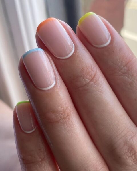 Zdjęcie dłoni z krótkimi, kwadratowymi paznokciami w kolorze nude, z french manicure w kolorach tęczy oraz reverse french manicure z białymi kreseczkami u nasady paznokci.