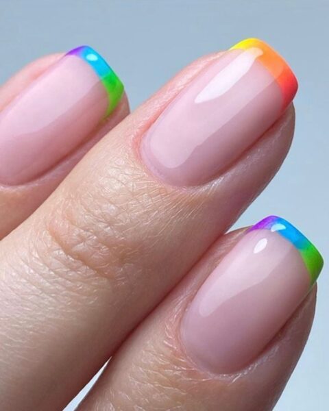 Zdjęcie dłoni ze zbliżeniem na kwadratowe paznokcie w stylu french manicure w kolorze tęczy z efektem ombre na brzegach.