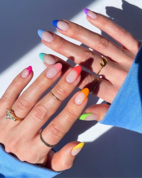 Zdjęcie dłoni z długimi, migdałowymi paznokciami z french manicure w kolorze tęczy. Każdy paznokieć ma inny kolor końcówki. Na palcach znajdują się liczne pierścionki.