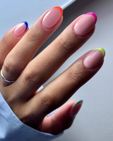 Zdjęcie dłoni ze zbliżeniem na długie, owalne paznokcie pomalowane na kolor nude i z kolorowymi końcówkami w stylu french manicure. Na palcu serdecznym znajduje się srebrna obrączka.