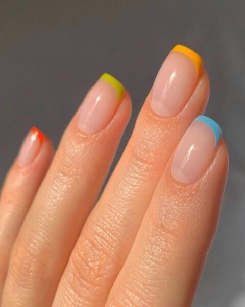 Zdjęcie dłoni ze zbliżeniem na kwadratowe paznokcie. Są pomalowane w stylu french manicure z tęczowymi końcówkami.