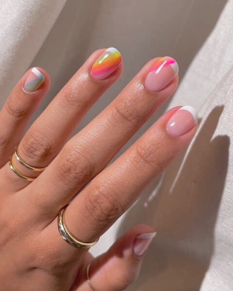 Zdjęcie dłoni ze zbliżeniem na owalne, średniej długości paznokcie z klasycznym french manicure i tęczową łuną na kilku paznokciach. Na palcach znajduje się kilka pierścionków. Dłoń pozuje na tle materiału w kolorze białym.
