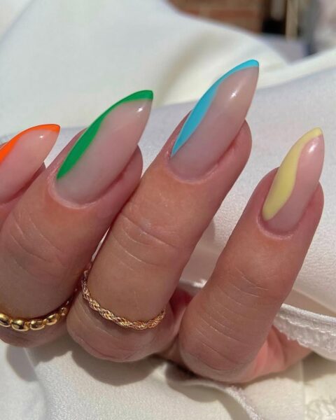 Zdjęcie dłoni z długimi, migdałowymi paznokciami w kolorze nude i ozdobione stylizacją wavy side french manicure z kolorowymi kreseczkami po bokach. Na palcach widać złote pierścionki. Dłoń pozuje na białym materiale.