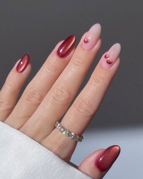 Zdjęcie dłoni z długimi, migdałowymi paznokciami. Dwa paznokcie mają kolor nude i wzorek wiśni, a pozostałe są wiśniowe z efektem glass nails. Na palcu wskazującym znajduje się pierścionek z cyrkoniami.