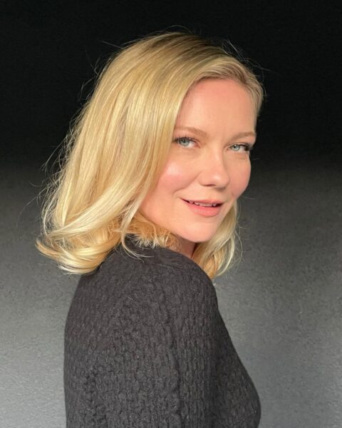 Zdjęcie aktorki Kirsten Dunst. Ma blond long boba do ramion lekko falowanego. Ubrana jest w szary sweter. Pozuje półbokiem i spogląda w bok, patrząc w obiektyw. Stoi na tle szarej ściany.
