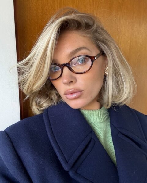 Zdjęcie modelki Elsy Hosk, która ma blond boba o dużej objętości. Ma lekko rozwiane, luźne włosy. Na nosie ma okulary korekcyjne. W uszach złote kolczyki. Ubrana jest w zielony sweter oraz granatowy płaszcz z kołnierzem. Pozuje na tle drewnianych drzwi.
