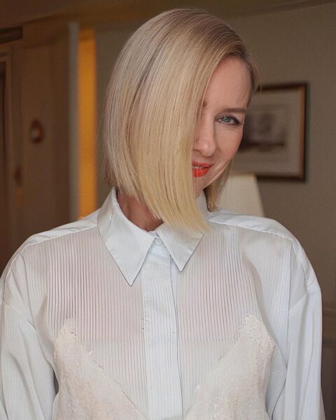 Zdjęcie aktorki Naomi Watts. Ma blond włosy ścięte na boba i uczesane w stylu sleek bob. Ubrana jest w białą koszulę. Włosy częściowo zasłaniają jej twarz. Pozuje w pokoju hotelowym.