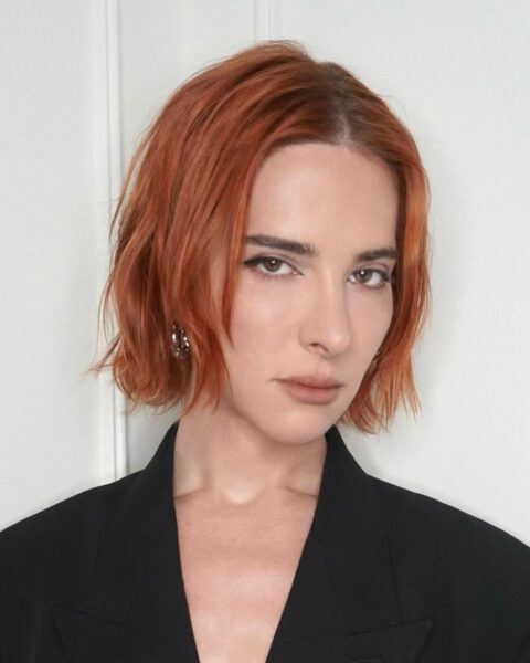 Zdjęcie modelki w fryzurze typu messy bob, czyli luźno, nonszalancko uczesanej. Włosy ma rude i bardzo krótkie. Ubrana jest w czarna marynarkę. W uszach znajdują się srebrne kolczyki. Pozuje na tle białej ściany.