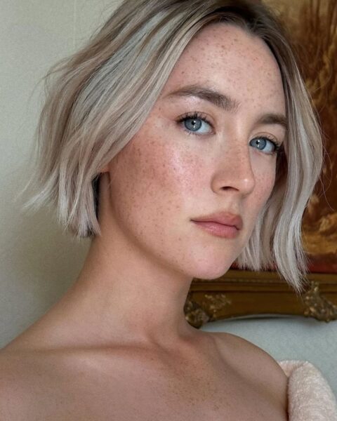 Zdjęcie aktorki Saoirse Ronan. To portret. Widać jej fryzurę typu bob w kolorze blond w stylu wavy bob. Ma naturalny makijaż z piegami na twarzy. Ma odsłonięte ramiona. Pozuje na tle ściany i widać za nią obraz w eleganckiej, zdobionej ramie.