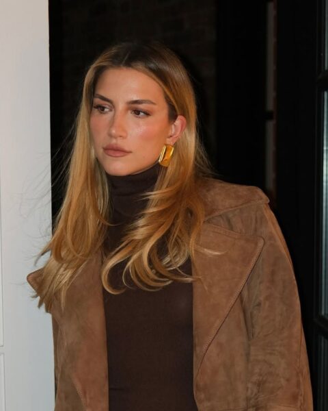 Zdjęcie influencerki Cass Dimicco. Stoi w drzwiach, ubrana jest w zamszowy, brązowy płaszcz, a także brązową torebkę . W ręce trzyma ciemnobrązową torebkę z motywem skóry krokodyla. Ma długie, rozpuszczone włosy w kolorze blond.
