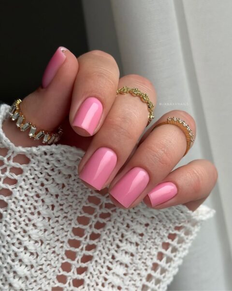 Zdjęcie dłoni z manicure na czerwiec 2025: paznokcie są długie, kwadratowe i pomalowane na cukierkowy, pastelowy różowy kolor. Na palcach widać kilka pierścionków. Widać też fragment rękawa ażurowego, białego swetra.
