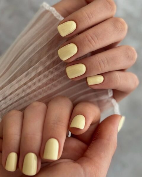 Zdjęcie dłoni ze zbliżęniem na długie, kwadratowe paznokcie w kolorze żółtym w odcieniu butter yellow. Dłonie pozują tak, by prezentować manicure. Widać fragment białego, tiulowego rękawa.
