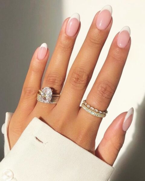 Zdjęcie dłoni ze zbliżeniem na długie, owalne paznokcie ozdobione wzorem french manicure. Na palcach widać liczne pierścionki. Widać też fragment kremowego rękawa bluzki z guzikami.
