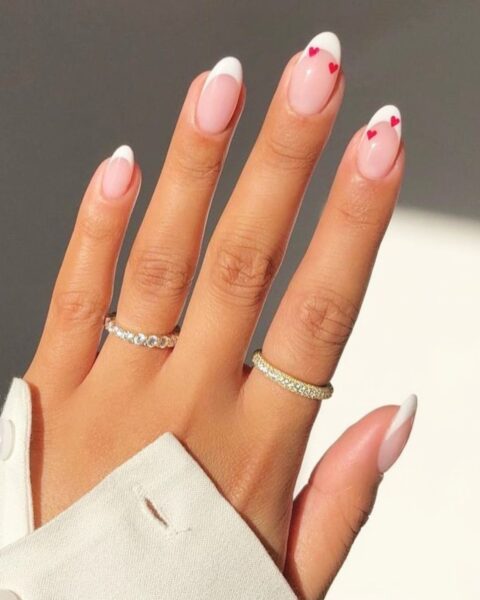 Zdjęcie dłoni ze zbliżeniem na długie, owalne paznokcie ozdobione wzorem french manicure oraz drobnymi, czerwonymi serduszkami. Na palcach znajdują się dwa pierścionki wysadzane cyrkoniami. Widać też fragment kremowego rękawa bluzki z guzikami.