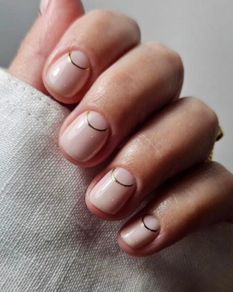 Zdjęcie dłoni ze zbliżeniem na krótkie, owalne paznokcie, które zdobi manicure śłubny ze złotym halfmoon na nude bazie.