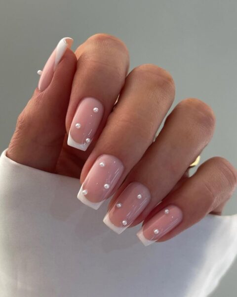 Zdjęcie dłoni ze zbliżeniem na długie, kwadratowe paznokcie ze wzorem french manicure oraz zdobieniem w postaci kilku drobnych perełek.
