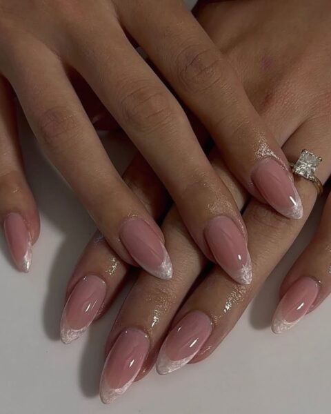 Zdjęcie dłoni ze zbliżeniem na paznokcie, długie i migdałowe, pomalowane w stylu french manicure z perłową końcówką. Na palcu serdecznym lewej dłoni znajduje się pierścionek z brylantem. Dłonie pozują ułożone jedna na drugiej, na białym blacie.
