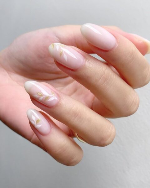 Zdjęcie dłoni ze zbliżeniem na długie, migdałowe paznokcie ze ślubnym manicure w kolurze nude i kwiatowym zdobieniem ze złotymi listkami.