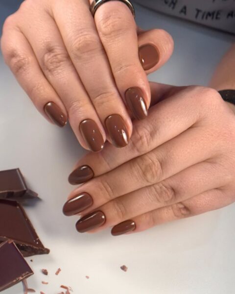 Zdjęcie dłoni, która ma długie, owalne paznokcie w kolorze brązowym o odcieniu mlecznej czekolady. Dłonie pozują na białym blacie i prezentują manicure. Obok leżą kawałki mlecznej czekolady.