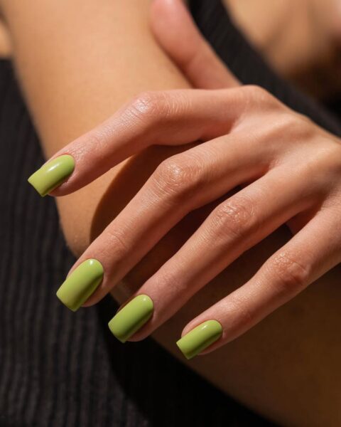 Zdjęcie dłoni ze zbliżeniem na manicure w kolorze limonkowej zieleni. Dłoń pozuje oparta o drugą rękę. Widać w tle czarny top kobiety.