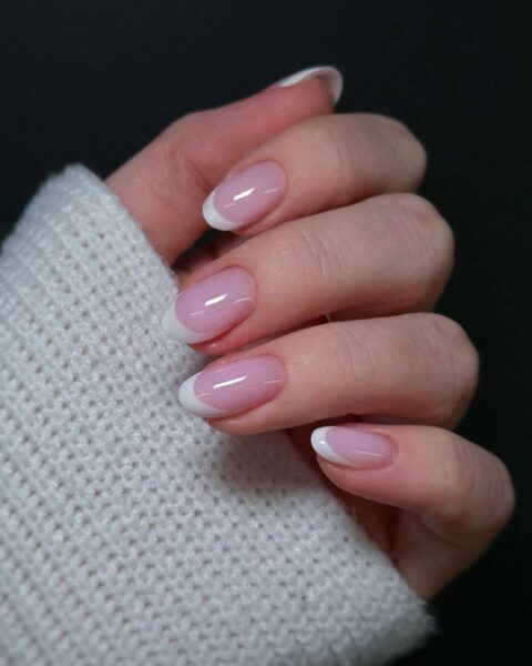 Zdjęcie dłoni z długimi, owalnymi paznokciami pomalowanymi w stylu french manicure. Dłoń pozuje na czarnym tle. Widać fragment rękawa kremowego swetra.