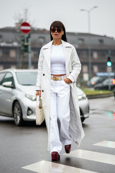 Kobieta w białych jeansach i białym crop topie z długim płaszczem w tonacji off-white, stylizacja uzupełniona szerokim paskiem Gucci, bordowymi platformami i białą torebką — miejski, letni look z pokazu mody ulicznej.