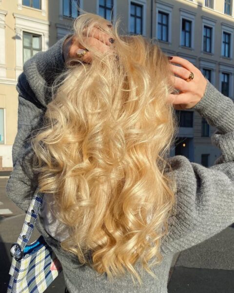 Zdjęcie kobiety z długimi, gęstymi blond włosami, które są lekko pofalowane. Pozuje z tyłu i widać całą fryzurę. Unosi ręce i przeczesuje włosy. Jest ubrana w szary sweter, czarne jeansy. Na ramieniu ma torebkę w niebieską kratkę. Idzie ulicą.