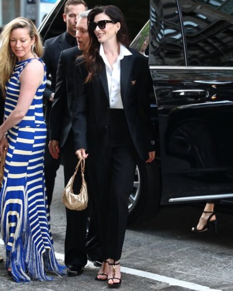 Zdjęcie Anne Hathaway. Aktorka ubrana jest w czarny garnitur, białą koszulę i czarne sandałki. Ma okulary przeciwsłoneczne na nosie oraz złotą torebkę w ręce. Długie włosy, ciemne i rozpuszczone. Wysiadła samochodu i zmierza na event.