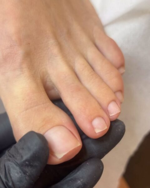 Zdjęcie stóp z niepomalowanymi, ale zadbanymi paznokciami. Pozują na łóżku do pedicure. Trzyma je dłoń kosmetyczki, która nosi czarną rękawiczkę.
