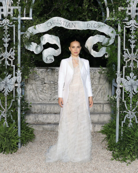 Jest to Natalie Portman na pokazie Dior Cruise 2026. Osoba ubrana w elegancką, długą suknię z warstwowego, półprzezroczystego materiału w odcieniu kości słoniowej, zestawioną z białym, dopasowanym płaszczem, stoi pod ozdobnym łukiem z napisem „Christian Dior”, otoczonym zielenią i metalowymi zdobieniami w stylu ogrodowym. Tło stanowi kamienny relief z postaciami historycznymi.