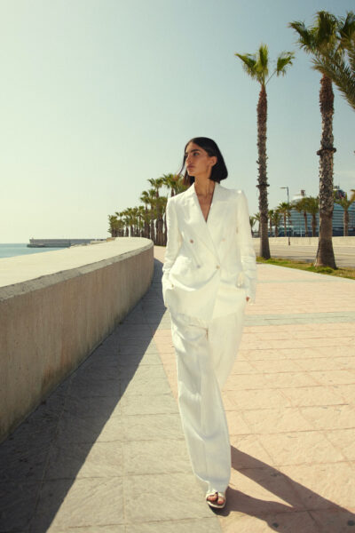 Kobieta w eleganckim, białym garniturze z podwójnym rzędem guzików, spacerująca wzdłuż nadmorskiej promenady z palmami i widokiem na port.