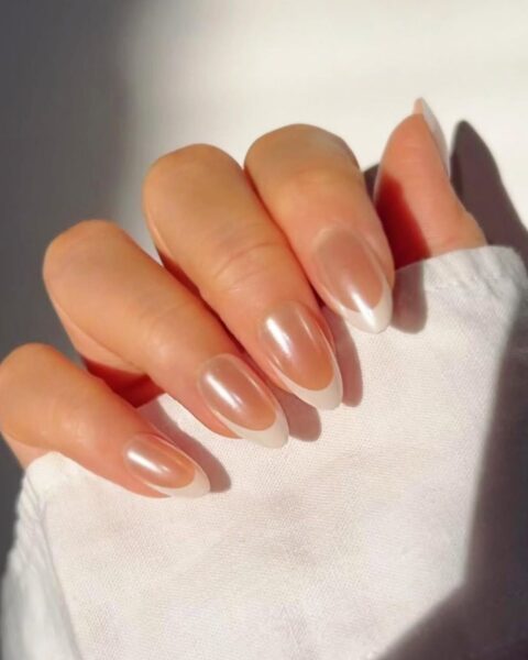 Zdjęcie dłoni i na długie, migdałowe paznokcie z french manicure wykończone perłowym wykończeniem. Dłoń układa się tak, by prezentować manicure. Widać fragment rękawa białej koszuli.