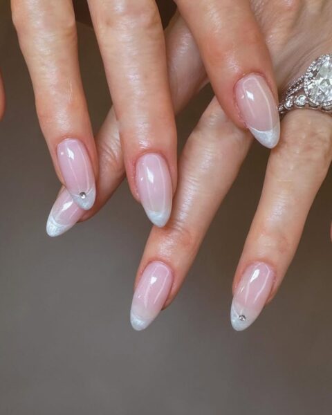 Zdjęcie dłoni i zbliżenie na długie, migdałowe paznokcie ze wzorem french manicure i dodatkowo ozdobione kryształkami. Na palcu serdecznym znajduje się masywny, srebrny pierścionek z brylantem.