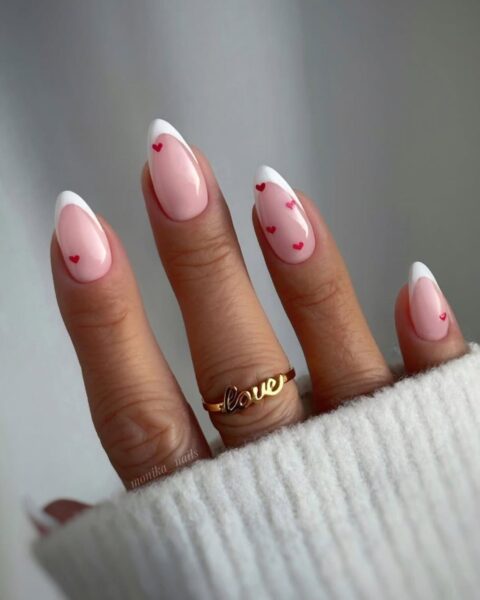 Zdjęcie dłoni ze zbliżeniem na długie, migdałowe paznokcie. Są ozdobione wzorem french manicure i dodatkowo ozdobione drobnym wzorem serduszek w kolorze czerwonym. Na środkowym palcu znajduje się złoty pierścionek.