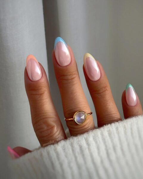 Zdjęcie dłoni ze zbliżeniem na długie, migdałowe paznokcie. Są ozdobione wzorem french manicure, ale w pastelowymi końcówkami i dodatkowo pokryte pyłkiem z chromowanym wykończeniem. Na palcu środkowym znajduje się złoty pierścionek z kamieniem.