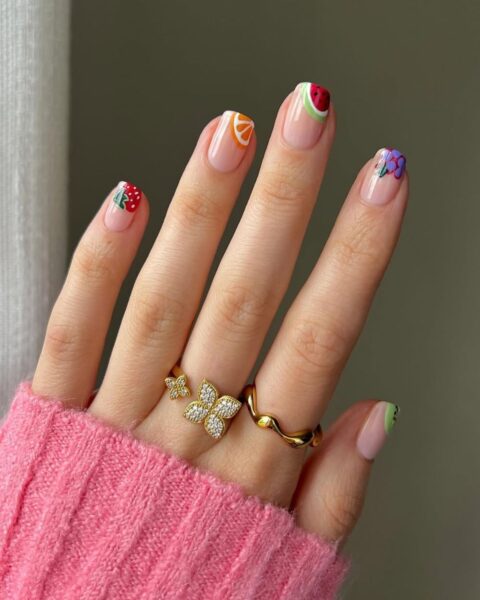 Zdjęcie dłoni z długimi, kwadratowymi paznokciami ze wzorem corner nail art. z owocowymi motywami. Na palcach znajdują się dwa złote pierścionki. Widać fragment rękawa różowego swetra.