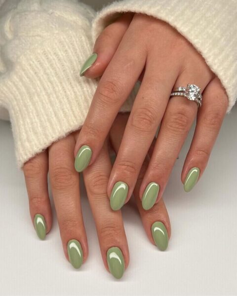 Zdjęcie dłoni ze zbliżeniem na długie, owalne paznokcie matcha nails, czyli w kolorze jasnej zieleni z mocnym połyskiem. Na palcu serdecznym znajduje się srebrny pierścionek z kryształem. Dłonie pozują na białym blacie i układają się tak, by prezentować manicure.