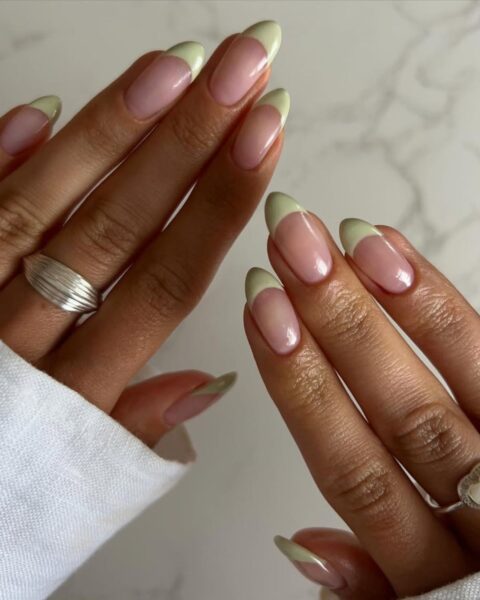 Zdjęcie dłoni z długimi, migdałowymi paznokciami z french manicure z zieloną końcówką zamiast białą. Na palcach widać srebrne pierścionki. Dłonie pozują na tle marmurkowego blatu.