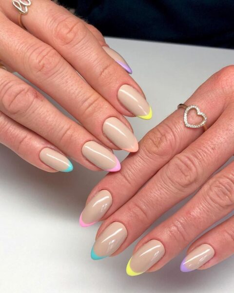 Zdjęcie dłoni z długimi, migdałowymi paznokciami pomalowanymi w stylu french manicure, ale z kolorowymi końcówkami. Na placach widać pierścionki. Dłonie pozują na tle białego blatu.