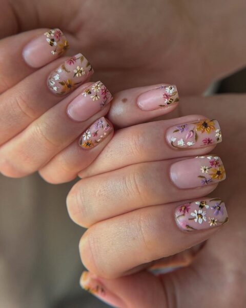 Zdjęcie dłoni z długimi, kwadratowymi paznokciami. Są ozdobione motywem kwiatów namalowanych w stylu polnych kwiatów techniką nail art. Dłonie pozują tak, by prezentować manicure.