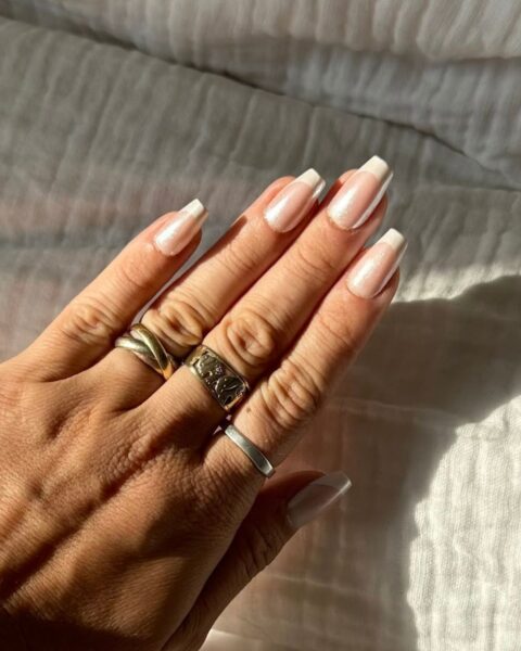 Zdjęcie dłoni ze zbliżeniem na długie, kwadratowe paznokcie pomalowane w stylu french manicure i pokryte perłowym topem. To manicure na wesele w maju. Na palcach znajduje się kilka pierścionków. Dłoń pozuje na tle białej pościeli muślinowej.