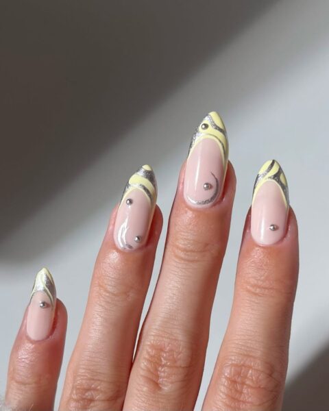 Zdjęcie dłoni ze zbliżeniem na długie paznokcie. Zdobi je wzór french manicure z żółtą, maślaną końcówką oraz abstrakcyjnymi, tribalowymi wzorami w kolorze srebrnym i z kryształkami.