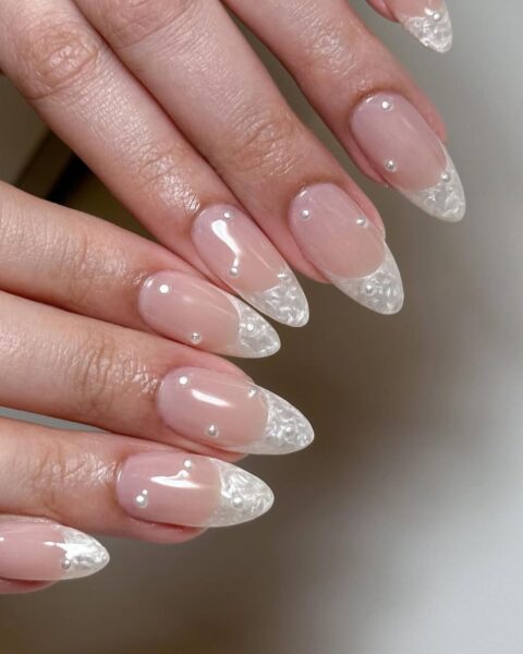Zdjęcie dłoni ze zbliżeniem na długie migdałowe paznokcie w stylu french manicure. Zamiast białych końcówek mają perłowe, a do tego zdobią je perełki 3D.