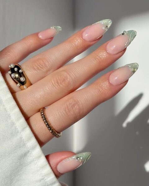 Zdjęcie dłoni ze zbliżeniem na długie migdałowe paznokcie. Zdobi je french manicure z jasnozieloną, perłową końcówką ozdobioną dodatkowo wzorem muszelek z żelu teksturującego oraz perełki 3D. Na palcach znajdują się dwa złote pierścionki.